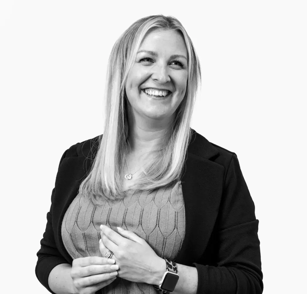 Smiling woman with long blonde hair wearing a sweater and blazer, standing against a plain white background.