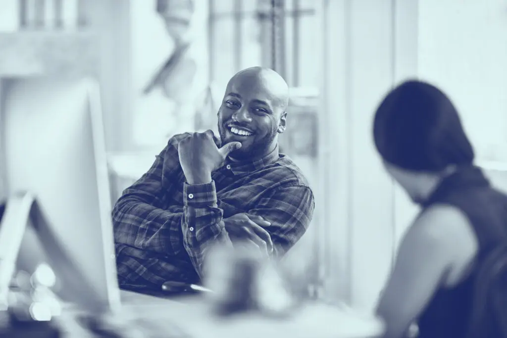 Smiling man sitting at a desk, chatting with a colleague in an office setting.