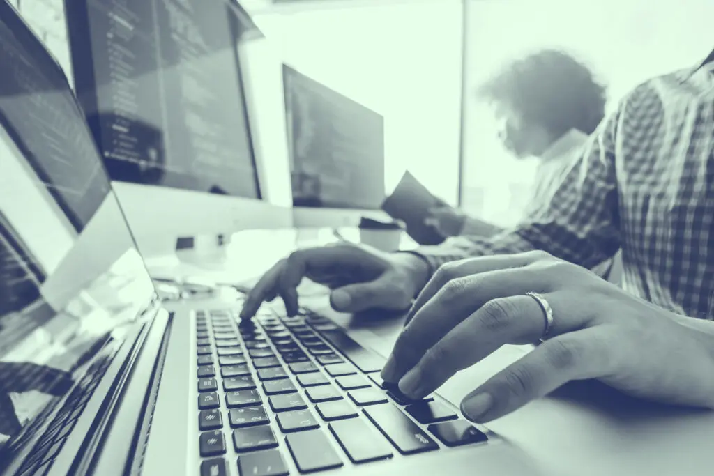 Close-up of hands typing on a laptop keyboard with computer monitors and people working in the background.