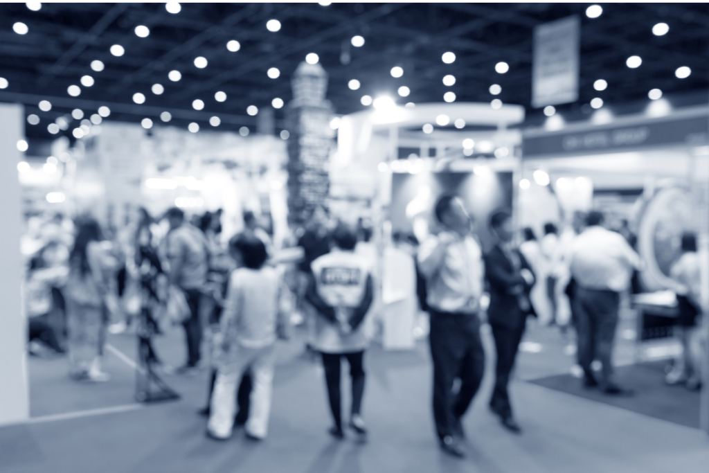 Blurry image of people walking around and talking at a busy indoor exhibition or trade show event.