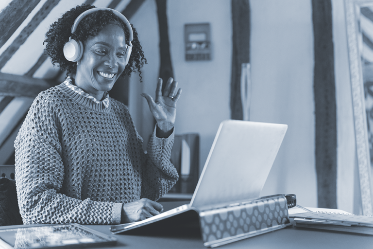 Smiling woman wearing headphones waves at laptop during a video call, sitting at a desk with books and papers.
