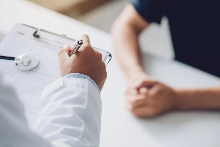 Doctor holding a clipboard and writing notes while talking to a patient with folded hands at a desk.