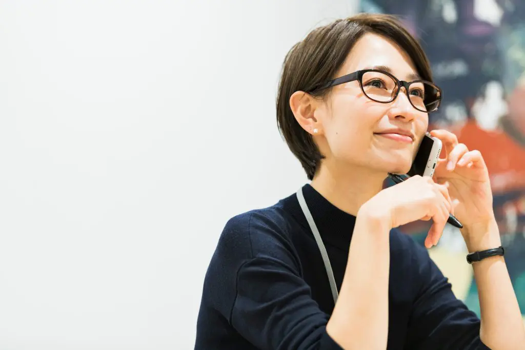 Smiling woman with glasses talking on a smartphone, wearing a dark top and lanyard.